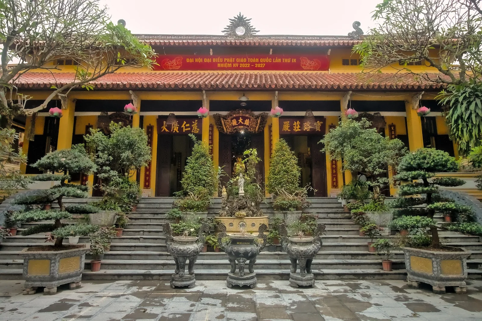 Yellow facade of Quan Su Temple in Hanoi with red-tiled roof, bonsai trees and stone steps leading to the entrance. Yellow facade of Quan Su Temple in Hanoi with red-tiled roof, bonsai trees and stone steps leading to the entrance.