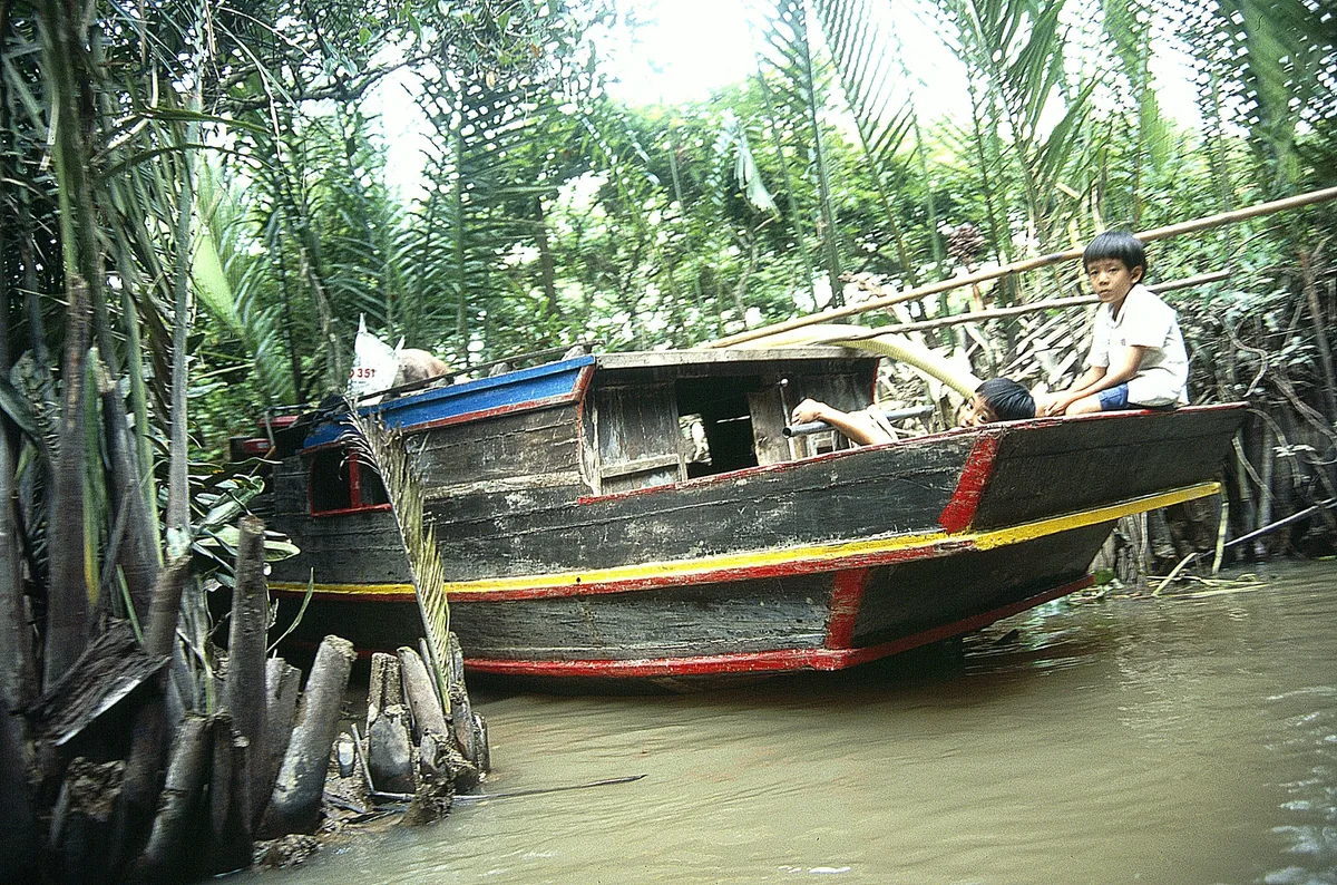 A wooden boat with children aboard rests on a narrow palm-lined canal in the Mekong Delta.